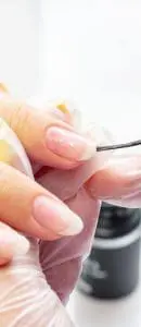 A close-up photograph shows a professional wearing translucent gloves carefully applying a clear base coat or gel to a client's natural fingernail.