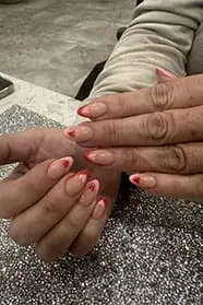 A close-up photograph features almond-shaped nails with a nude base, finished with red and pink French tips that are shaped into delicate hearts at the center.