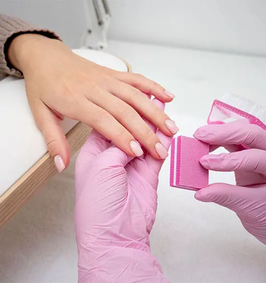 A close-up photograph shows a nail technician wearing pink gloves using a pink buffing block to smooth a client's short, square-shaped nails finished in a pale pink polish.