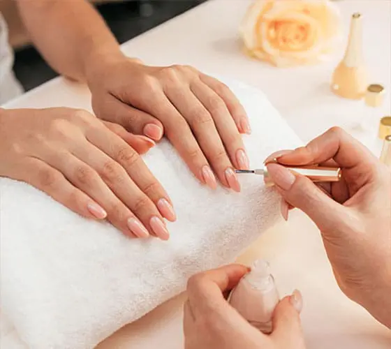 A close-up photograph shows a nail technician's hands carefully applying a sheer, pale pink polish to a client's long, almond-shaped fingernails.