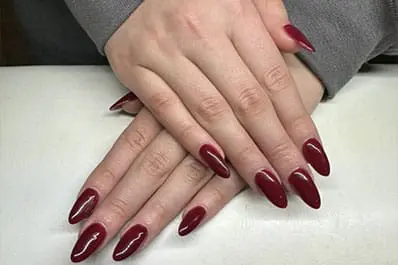 A close-up photograph displays a person's hands crossed over a white surface, featuring long, almond-shaped nails painted in a glossy, deep burgundy finish.
