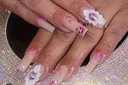 A close-up photograph showcases long, square-shaped nails featuring pink ombré tips, a small pumpkin illustration, and 3D white floral charms with iridescent centers.