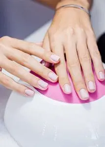 A close-up photograph shows a person’s hands featuring a clean, square-shaped manicure in a soft, matte lavender-pink shade, resting on a white and pink LED nail lamp.