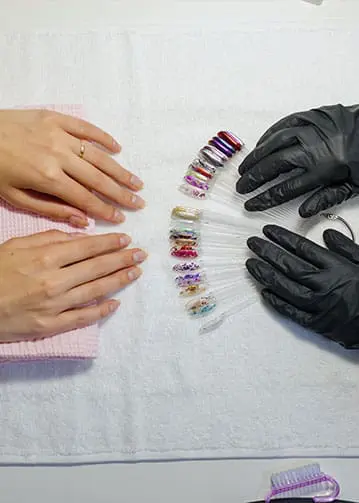 A professional nail technician wearing black gloves presents a fan of decorative nail swatches, featuring various metallic and glitter finishes, to a client with natural nails.