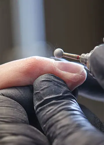 A close-up photograph captures a nail technician wearing black gloves as they use a spherical drill bit on an electric file to precisely refine a client's cuticle.