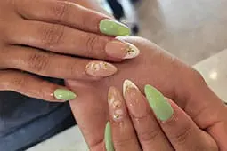 A close-up photograph showcases almond-shaped nails with a light green and neutral palette, featuring solid green polish, white French tips with gold accents, and delicate white floral designs.