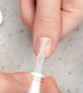 A close-up photograph shows a person's thumb receiving a coat of clear, glossy nail polish or base coat applied with a small brush.