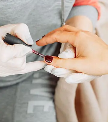 A close-up photograph captures a technician wearing white gloves carefully applying a deep burgundy polish to a client's fingernails.