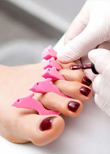A close-up photograph shows a technician wearing white gloves applying dark burgundy polish to a client's toenails, which are separated by pink, dolphin-shaped foam toe separators.