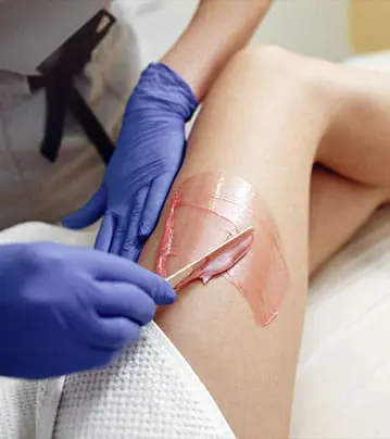 A close-up photograph shows a professional technician wearing blue gloves using a wooden spatula to apply a layer of pink wax to a client's leg.