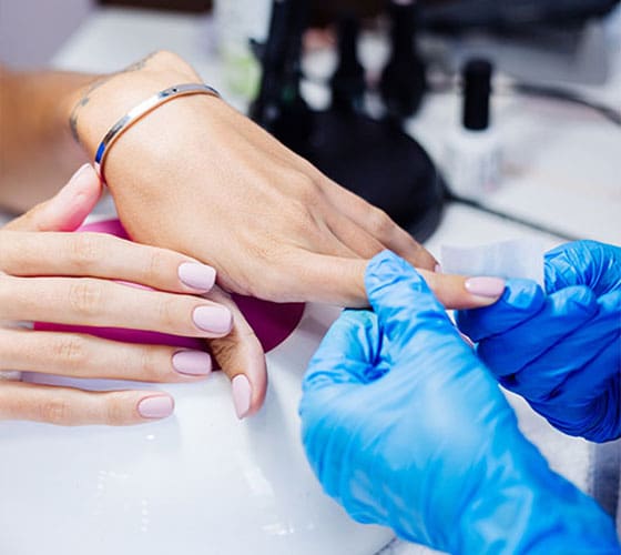 A professional wearing blue gloves carefully wipes a client's freshly cured, pale pink square-shaped nails after they have been placed under a white and pink LED lamp.