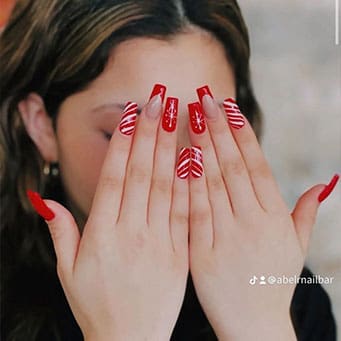 A person covers their face with hands featuring long, square-shaped red nails decorated with white peppermint swirls and sparkling starburst patterns.