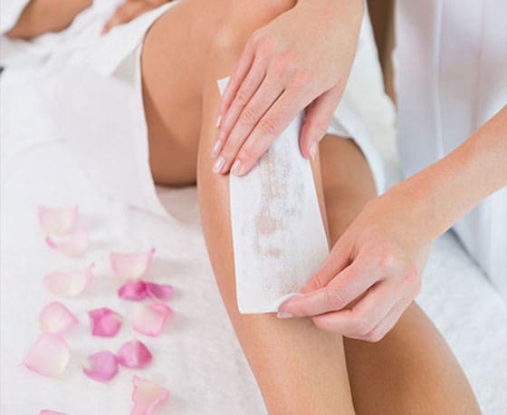 A close-up photograph shows a professional technician applying a white waxing strip to a client's leg to remove hair.