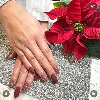 A high-angle photograph shows hands with almond-shaped nails featuring a combination of matte burgundy and shimmering gold-flecked polish, posed next to a vibrant red poinsettia flower.