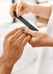 A close-up photograph shows a person using a black emery board to carefully shape and smooth a client's fingernails, which are finished in a neutral taupe polish.