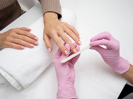 A close-up photograph shows a nail technician wearing pink gloves carefully shaping a client's fingernails with a professional nail file.