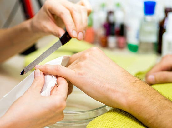 A professional nail technician uses a metal nail file to carefully shape the edge of a client's natural fingernail during a manicure service.