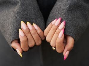 A close-up photograph features a person's almond-shaped fingernails with a French manicure, showcasing vibrant yellow tips on one hand and bright pink tips on the other.