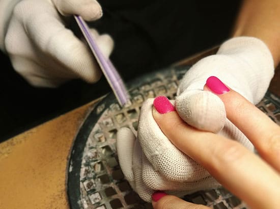 A professional nail technician wearing white textured gloves uses a purple and white file to shape the edge of a client's bright pink manicure over a salon ventilation grate.