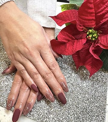 A high-angle photograph shows hands with almond-shaped nails featuring a combination of matte burgundy and shimmering gold-flecked polish, posed next to a vibrant red poinsettia flower.