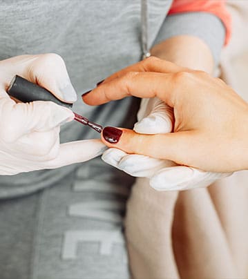 A close-up photograph captures a technician wearing white gloves carefully applying a deep burgundy polish to a client's fingernails.