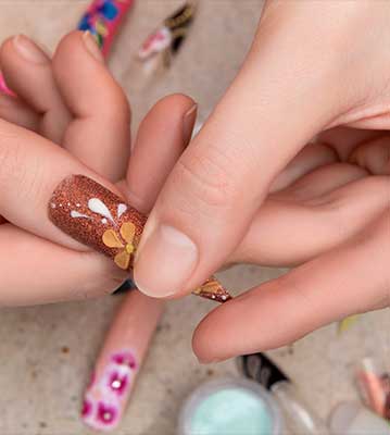 A close-up photograph shows a person's hands holding an artificial nail tip featuring a bronze glitter base decorated with a delicate orange and white floral design.