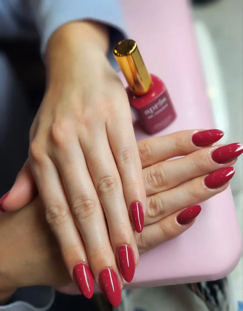 A close-up of hands with almond-shaped nails painted in a deep, glossy crimson red. One hand rests over the other near a bottle of red nail polish on a pink surface.