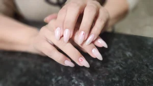 A pair of hands resting on a dark surface showcasing long almond nails with an iridescent pearlescent pink finish and tiny heart or gem accents on the ring fingers.