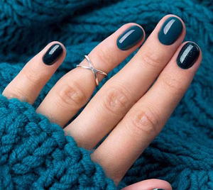 Hand with a silver "X" ring and glossy teal-blue manicured nails resting on a blue knit sweater.